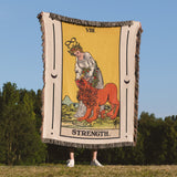 Person holding a 'Strength' tarot card blanket outdoors with trees and blue sky in the background