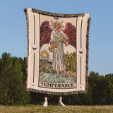 Person holding a 'Temperance' tarot card blanket outdoors with trees and blue sky in the background