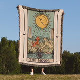 Person holding a 'The Moon' tarot card-style blanket outdoors with trees and blue sky in the background.