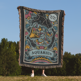 Person holding a colorful Aquarius zodiac-themed blanket outdoors with trees and blue sky in the background