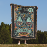 Person holding a colorful 'Libra' zodiac-themed blanket outdoors with trees and blue sky in the background.