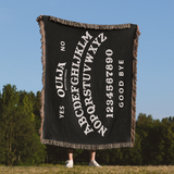 Black Ouija board-themed blanket held up against a clear blue sky with trees in the background